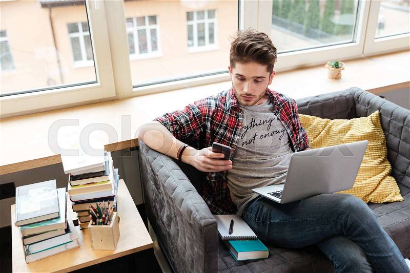 Surprised man looking at phone, holding laptop on kness and sitting on sofa in office. Top view, stock photo
