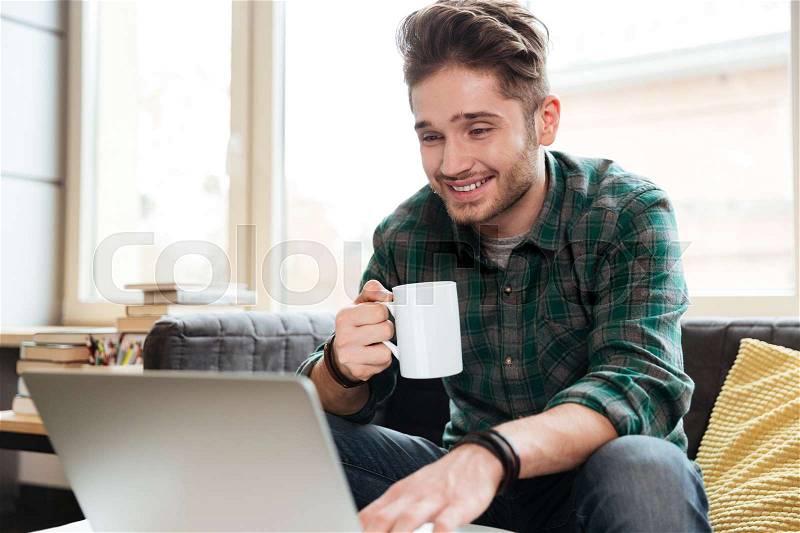 Happy man in green shirt sitting on sofa with cup of tea and looking at laptop. Coworking, stock photo