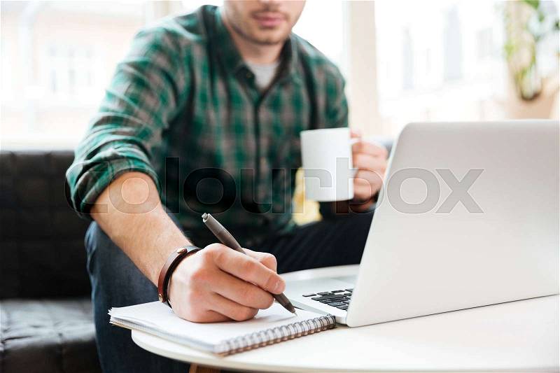 Cropped image of man in green shirt sitting at the table with laptop, holding cup of tea and writing something on notebook, stock photo