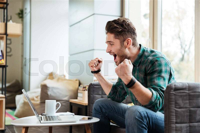 Side view of happy man sitting on sofa by the table with laptop. Man with open mouth. Cowerking, stock photo