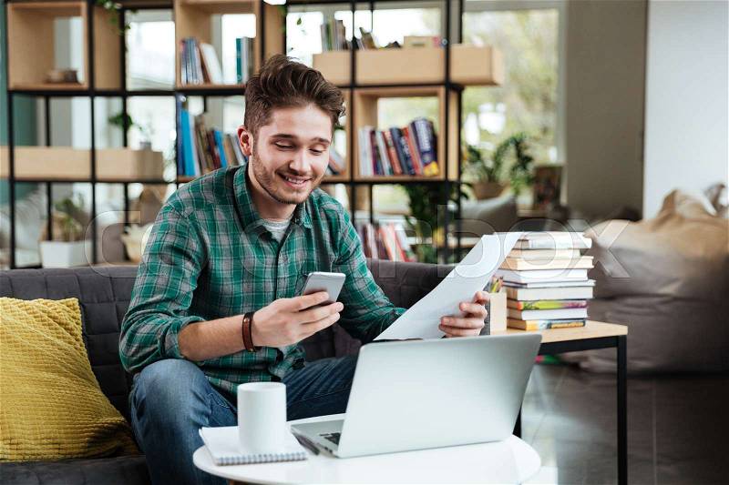 Happy man in green shirt with documents looking at phone and sitting on sofa by the table with laptop. Coworking, stock photo