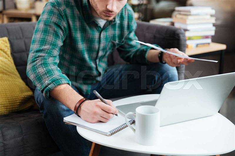 Cropped image of man in green shirt sitting on sofa with laptop and documents and writing by the table. Coworking, stock photo