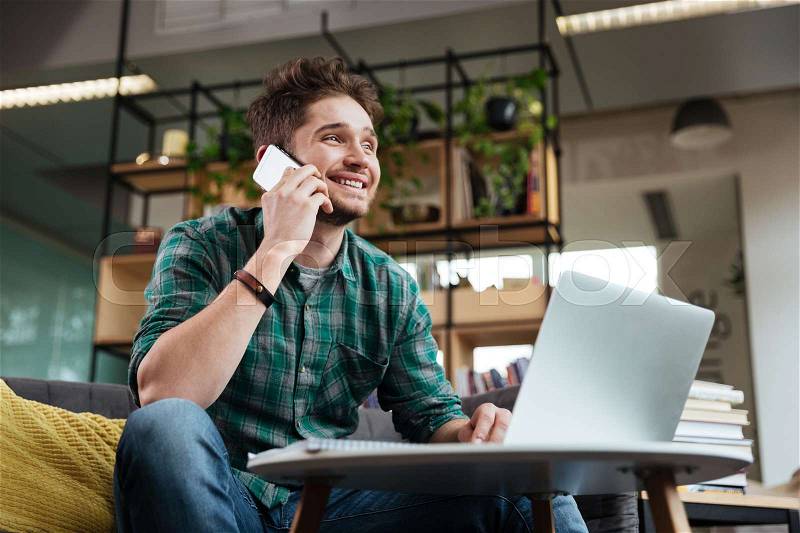 Happy man in green shirt sitting on sofa by the table with laptop and talking on phone in office. Coworking, stock photo