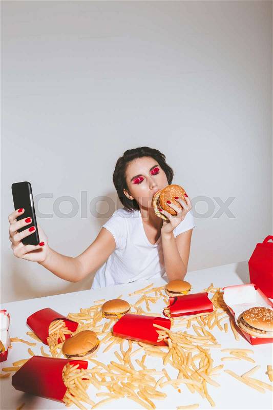 Portrait of attractive young woman eating burger and taking selfie with cell phone over white background, stock photo