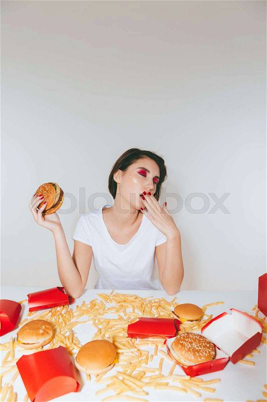 Tired young woman eating burger and yawning at the table over white background, stock photo