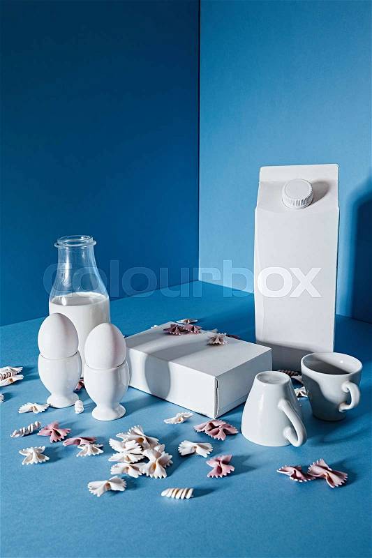 Bottle of milk, two blank boxes, white coffee cups and pasta bows over blue background, stock photo