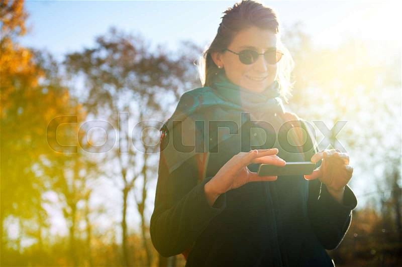 Gorgeous beautiful young caucasian woman messaging on the futuristic smart-phone device in the park outdoor background, chatting, using internet, soicial network, photo sharing services and messengers with beautiful sun flare behind her, stock photo