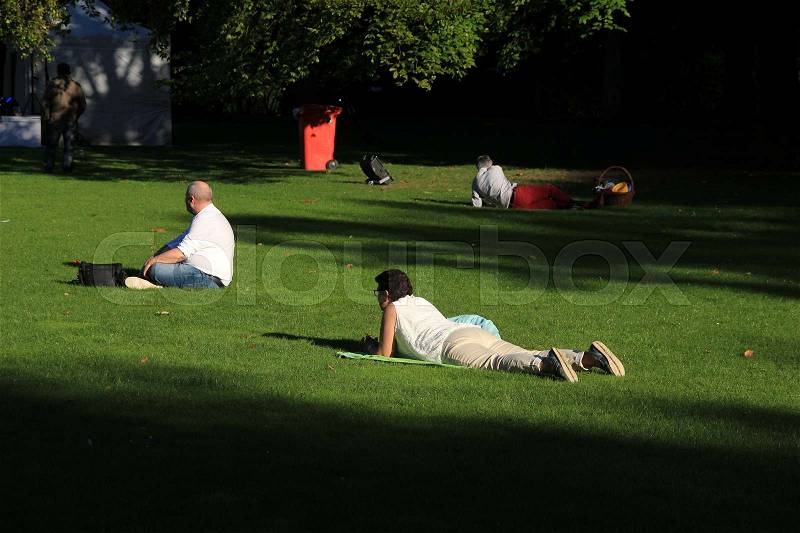 The separate people, men and wife are sitting or lying in the grass with sun and shadow sides in the urban park in the city in the summer, stock photo
