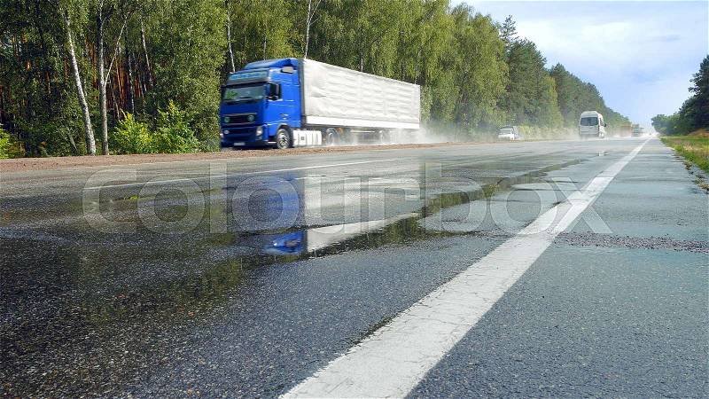 View from ground of traffic on wet road, highway with mist splash after rain with green trees on a roadside on background. Trucks are passing by camera in perspective, stock photo