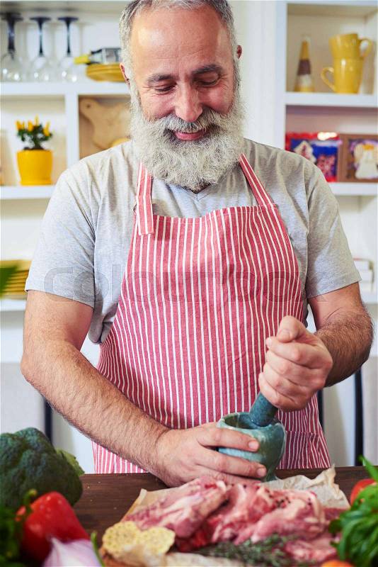 Mid shot of a man grinding spices on a cutting board. He wears stripped red and white apron standing isolated in the kitchen. Trying to prepare a nutritious dinner , stock photo