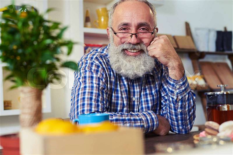 Close-up of happy senior man looking at the camera. Sitting at the table and leaning his head on the hand. Side rack focus on mature, bearded man , stock photo
