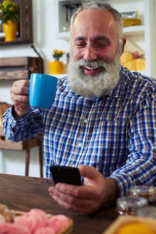 Close-up of mature man enjoying a cup of tea. Sitting at the table in the kitchen. Texting via smartphone. Wearing checked shirt , stock photo