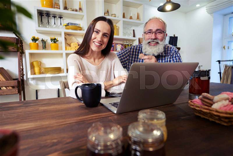 Close-up of cheerful couple. People sitting in the kitchen, looking at the camera. Using laptop and working in the morning. Delicious sweets on the wooden table, stock photo