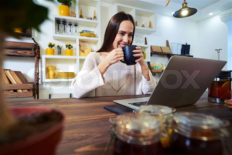 Low angle of delighted woman. Having a cup of tea while looking at the screen of the laptop. Sitting in the kitchen. Wearing a warm sweater , stock photo