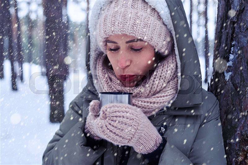 Close-up of pretty female blowing on tea while enjoying a mug of tea. Winter fashion. Portrait of a beautiful woman in warm clothes outdoor nearby the tree, stock photo
