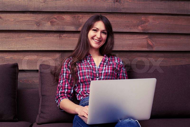 Modern youth using silver laptop while sitting intdoors on the sofa over wooden background. Wearing casual clothes. Working at home, stock photo