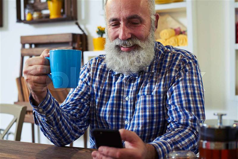 Side clos-up of positive man sitting at the kitchen table. Drinking a cup of tea, looking at the screen of the smartphone. Kitchen interior made in wooden style , stock photo