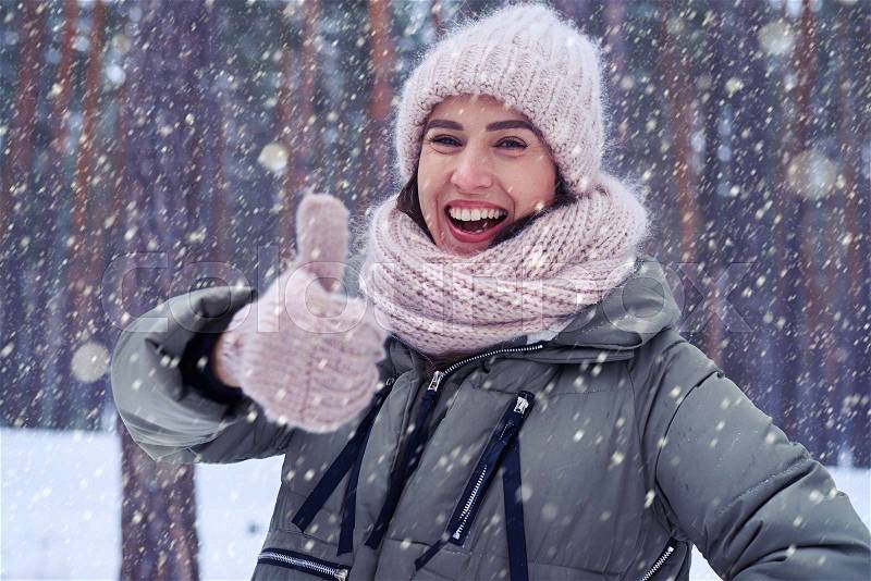 Close-up portrait of elated woman holding thumb up and smiling. Wearing warm pink knitted cap and scarf. Winter concept, stock photo