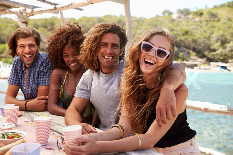Four friends sitting at a table by the sea look to camera, stock photo