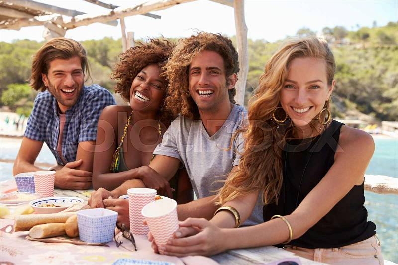 Four friends sitting at a table by the sea look to camera, stock photo