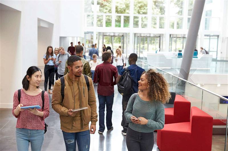Students holding tablets and phone talk in university lobby, stock photo