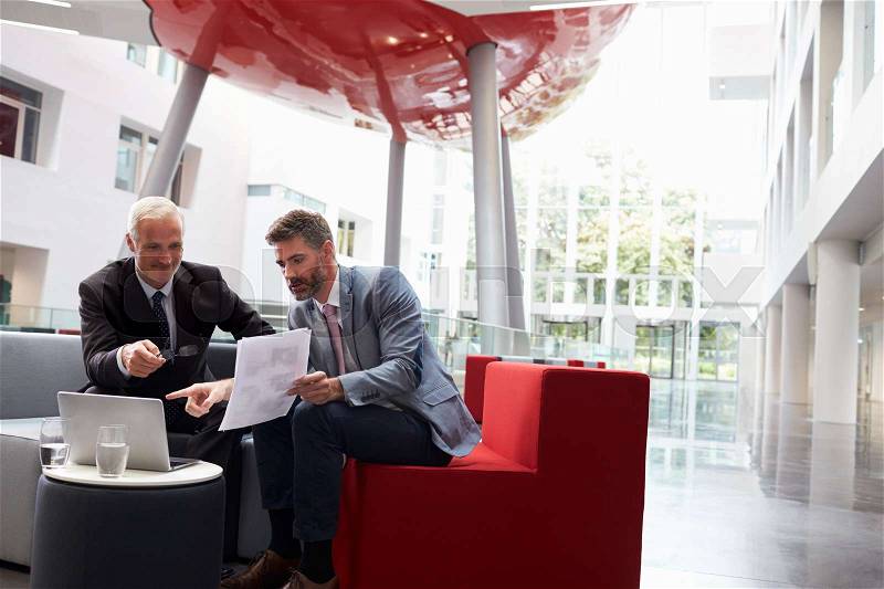 Two Businessmen Discuss Document In Lobby Of Modern Office, stock photo