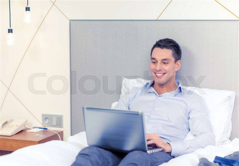 Business, technology, internet and hotel concept - happy businessman with laptop computer lying in hotel in bed, stock photo