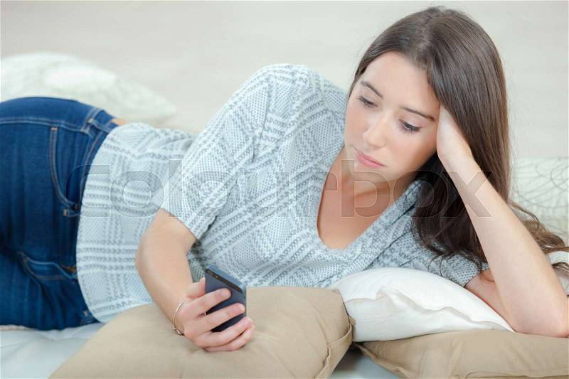 Lady lying on couch looking at cellphone, sad expression, stock photo