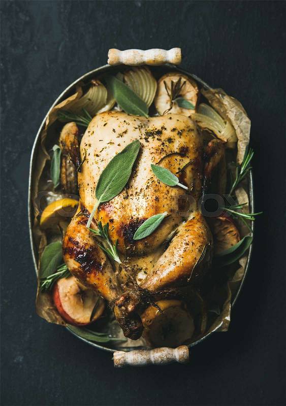 Oven roasted whole chicken with onion, apples and sage in metal serving tray over dark stone background, top view, selective focus. Celebration food concept, stock photo