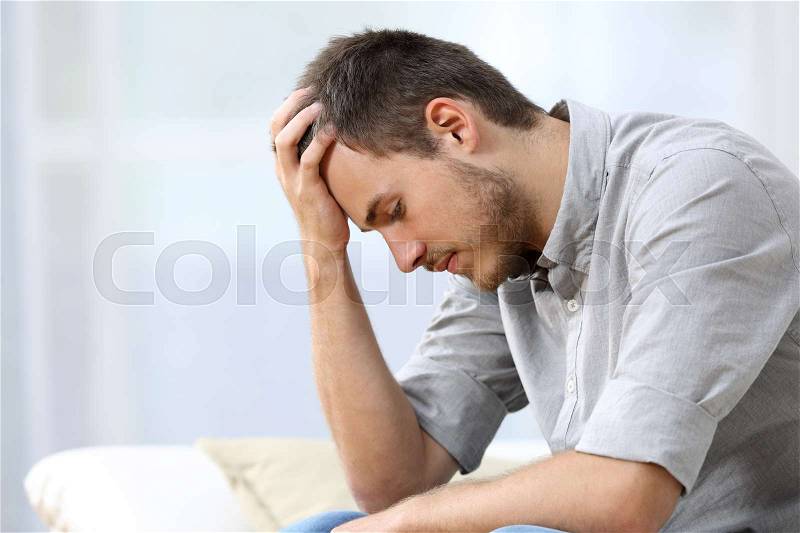Side view of a sad man with a hand on the head sitting on a couch in the living room at home, stock photo
