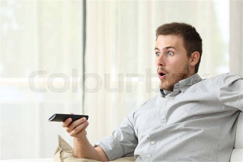 Surprised man zapping watching tv sitting on a couch in the livingroom at home, stock photo