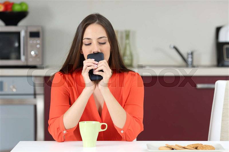 Doubtful woman looking down and wondering if she must call on the phone or waiting for a message in the kitchen at breakfast, stock photo