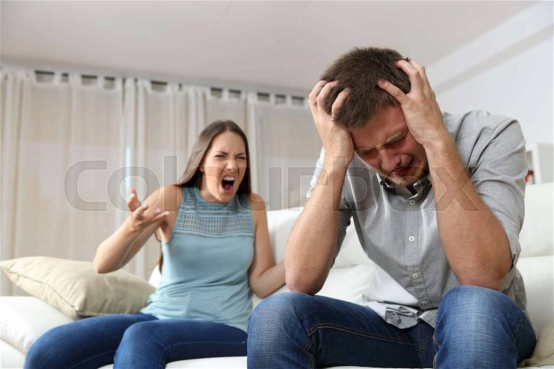 Couple arguing. Wife shouting to her desperate husband sitting on a couch in the living room at home, stock photo