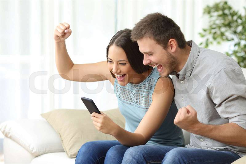 Excited couple watching media content together using a mobile phone sitting on a couch in the living room of a house, stock photo