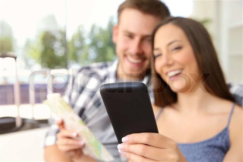 Couple of tourists searching location in a phone during vacations in an hotel or apartment with a window in the background, stock photo