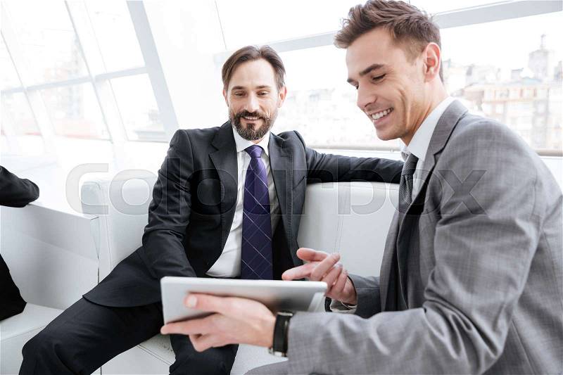 Two business partners talking in office, sitting on sofa and looking at tablet computer, stock photo