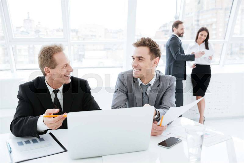 Laughing Business partners sitting by the table with laptop and documents with colleagues on background, stock photo