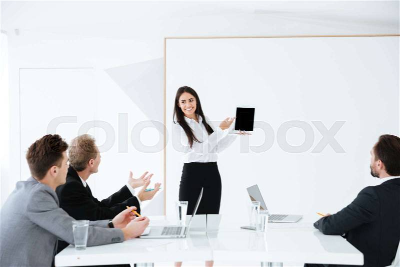 Woman showing blank tablet computer screen and standing near the board on conference with her colleagues which sitting by the table, stock photo