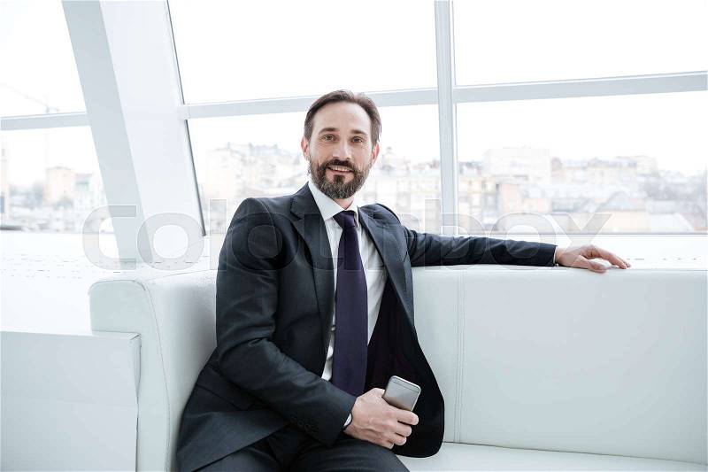 Elderly bearded business man sitting on sofa with phone near the window in office, stock photo