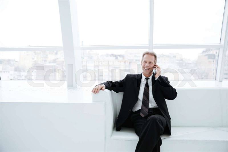 Elderly business man talking on phone and sitting on sofa near the window in office, stock photo