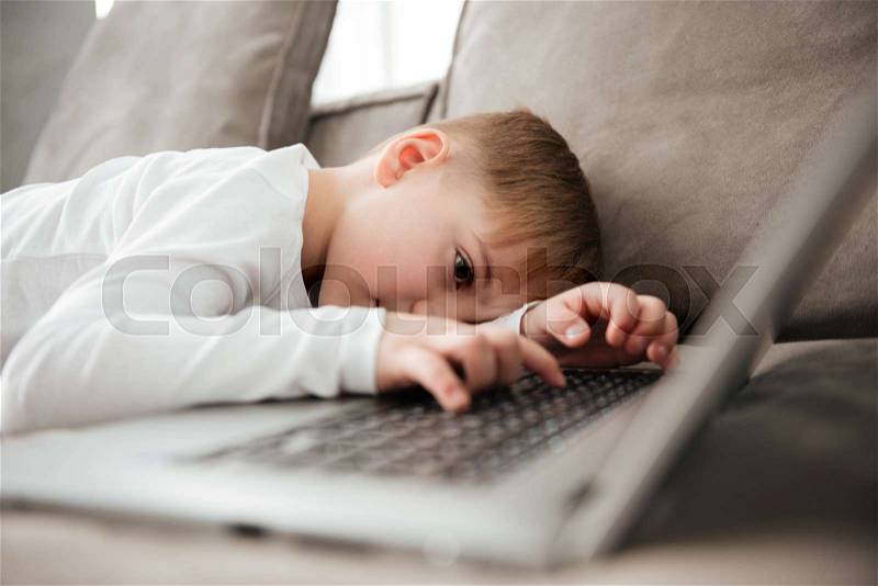 Picture of little alone sad boy lies on sofa while using laptop computer, stock photo