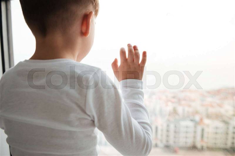 Picture of alone sad little boy near window waiting for parents at home. Look at window, stock photo