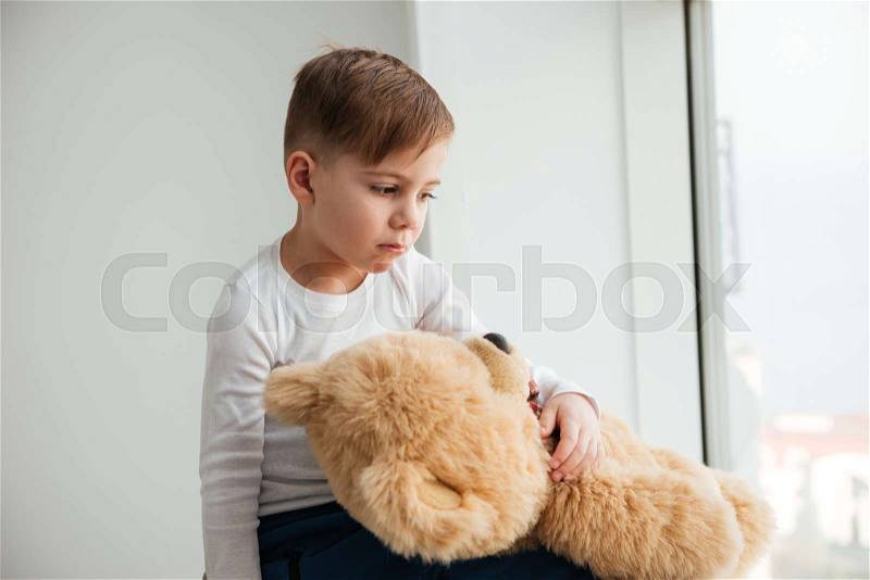 Photo of alone sad little boy near window with teddy bear waiting for parents at home. Look aside, stock photo