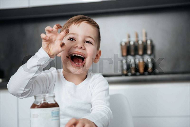 Picture of happy boy standing in the kitchen while eating sweeties. Look aside, stock photo