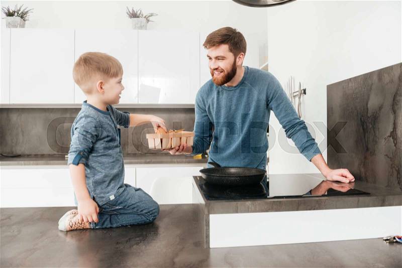 Photo of bearded father dressed in blue sweater cooking at kitchen with his little cute son, stock photo