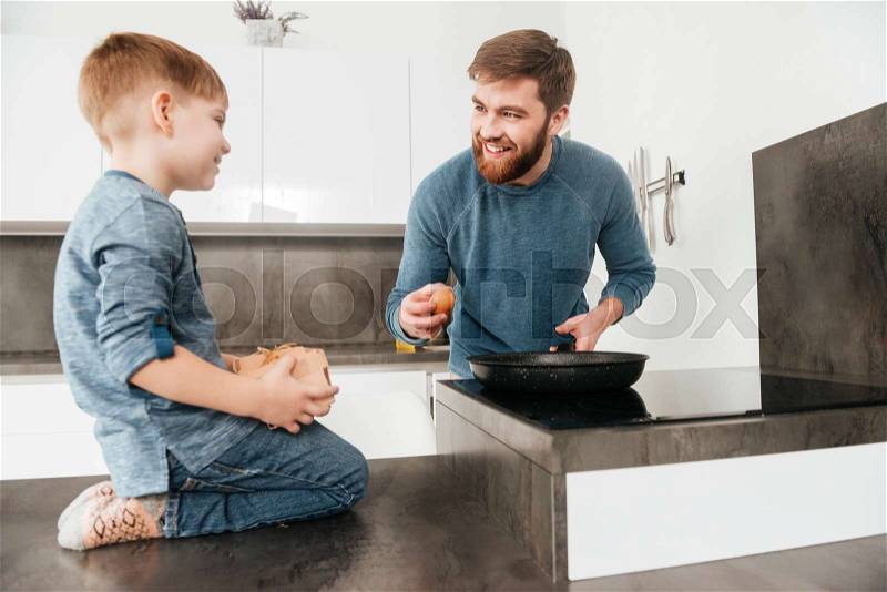 Picture of happy bearded father dressed in blue sweater cooking at kitchen with his little cute son, stock photo