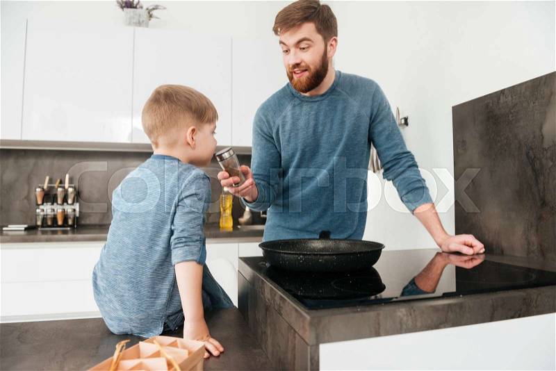 Image of bearded father dressed in blue sweater cooking at kitchen with his little cute son, stock photo