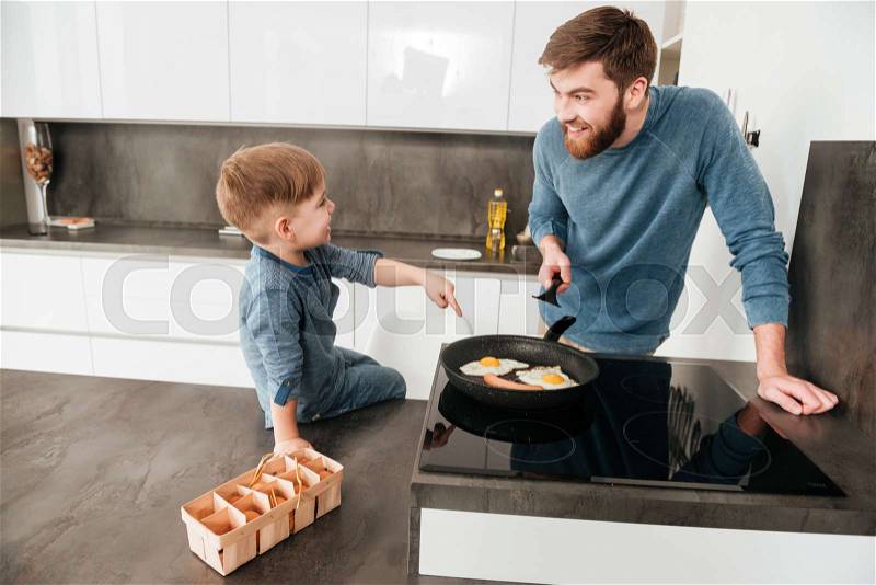 Photo of handsome bearded father dressed in blue sweater cooking at kitchen with his little cute son, stock photo