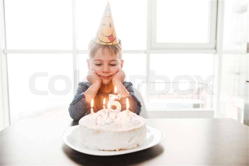 Photo of dreaming birthday boy sitting in kitchen near cake while smiling. Look at cake, stock photo