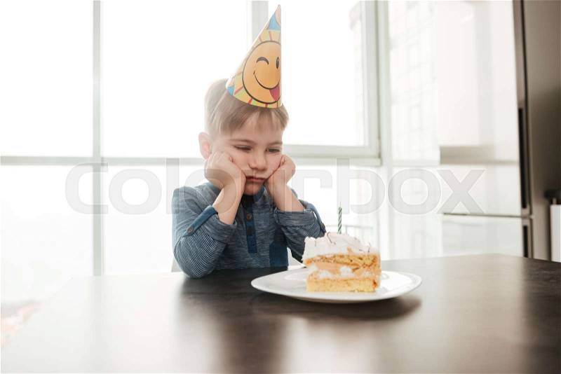Photo of sad birthday boy sitting in kitchen near cake alone. Look at cake, stock photo